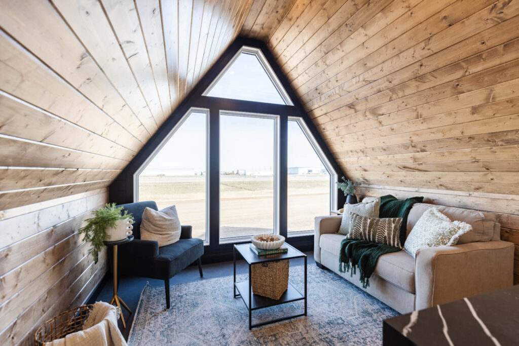 An a-frame tiny home with large black framed windows, wood accents, and cozy seating in the living area