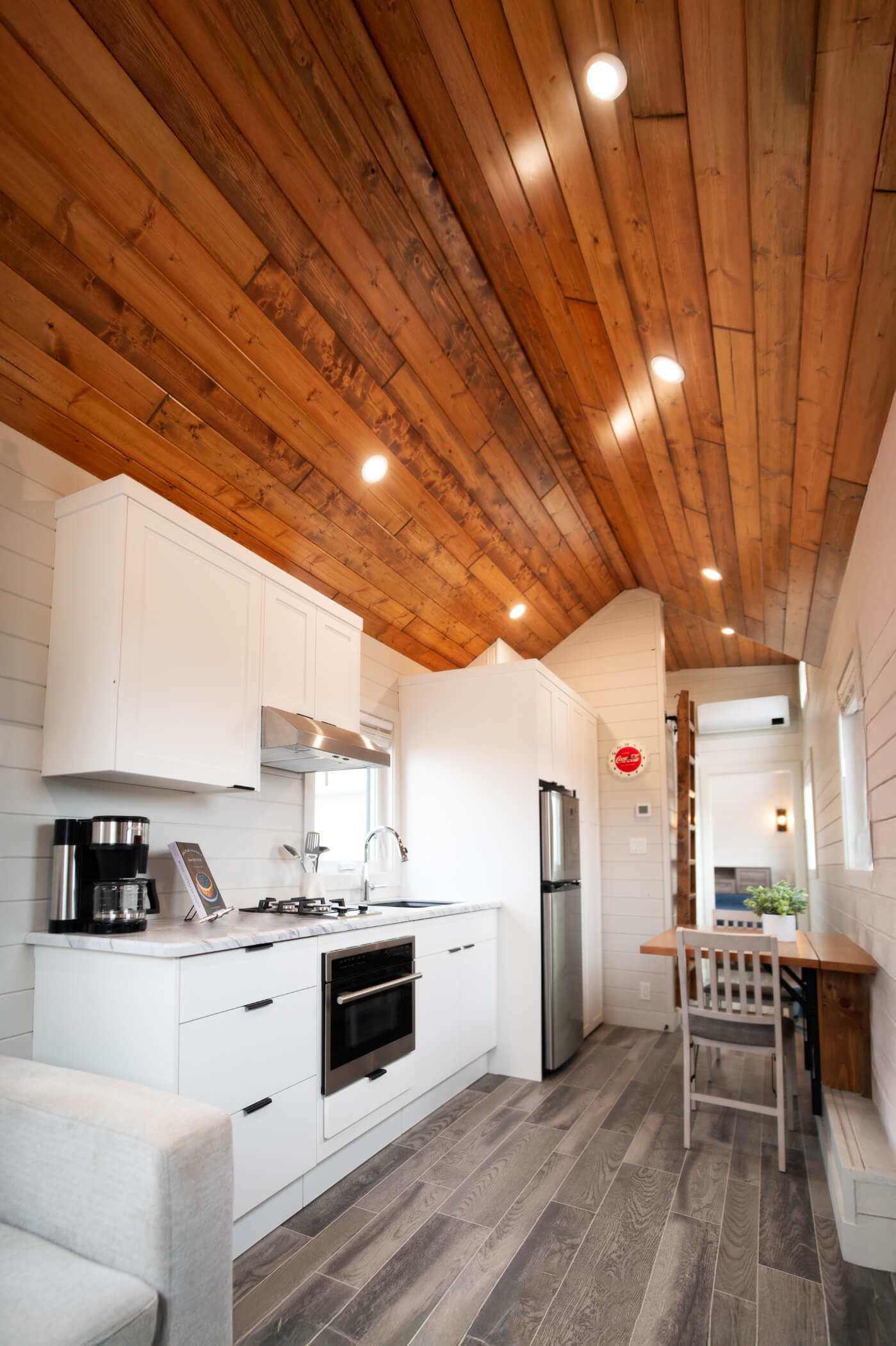 Bright interior of an Amberwood Tiny Home showcasing wood ceilings, white kitchen cabinetry, stainless appliances, and a compact dining area.