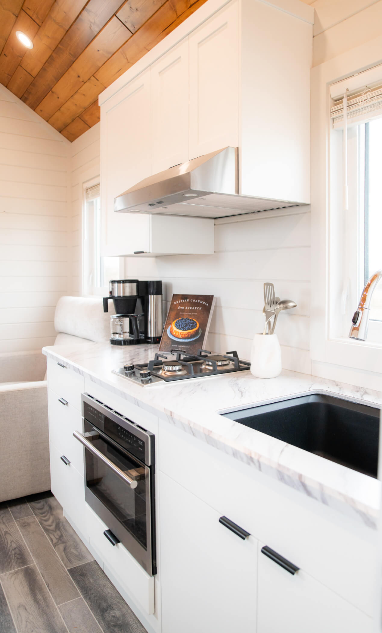 Bright modern kitchen inside an Amberwood Tiny Home featuring white cabinetry, gas cooktop, stainless oven, and marble-style countertops.