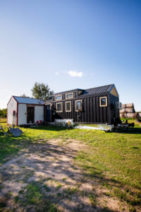Black tiny home on trailer parked on rural property beside small shed and hay bales